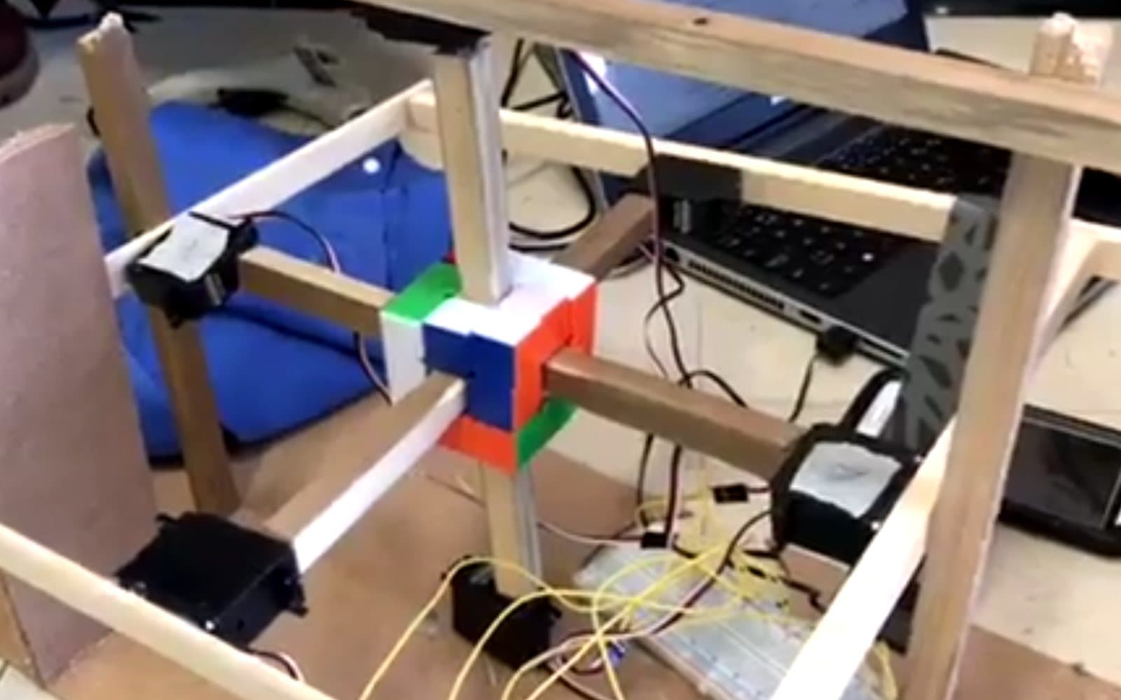 A wooden frame holding a Rubik's cube, with stepper motors on each axis, wired to a breadboard and a laptop